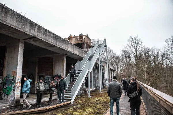 Afbeelding van meerdere mensen die lopen of stilstaan op een vlonderroute in een natuurgebied rondom een verlaten gebouw